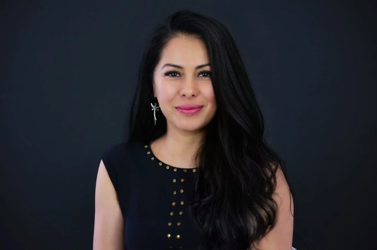 Retrato de una mujer joven con cabello largo y oscuro, vestido negro y aretes en forma de cruz, frente a un fondo negro.