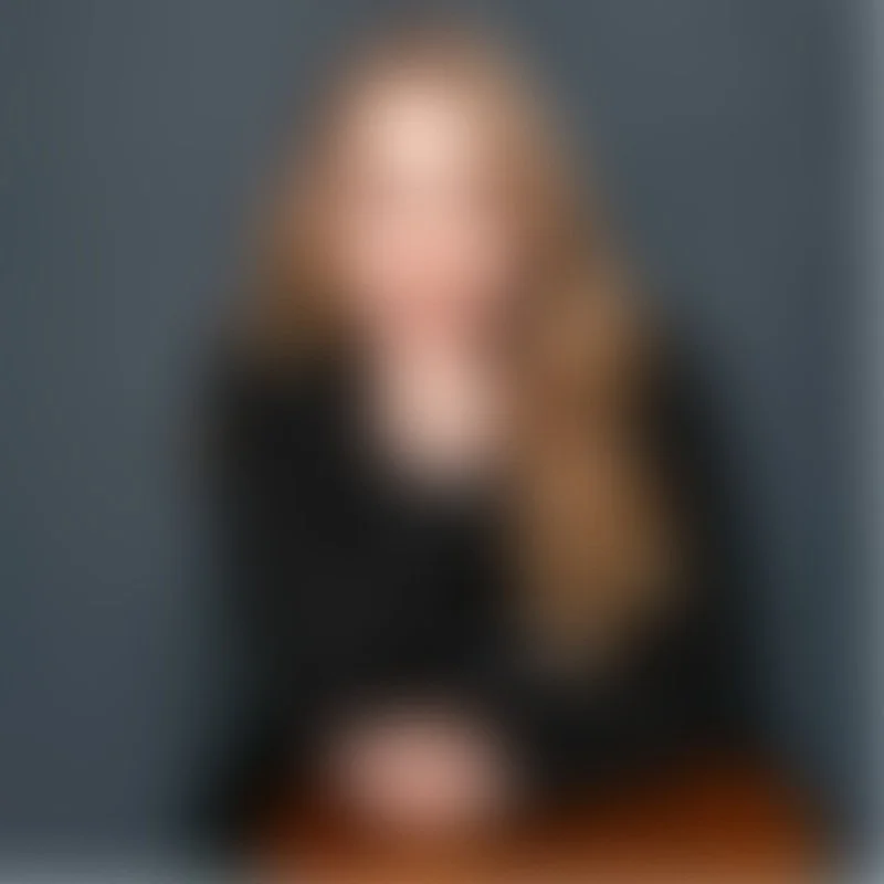 Imagen borrosa de una mujer con cabello rubio, usando ropa negra, sentada frente a una mesa, en un fondo gris.