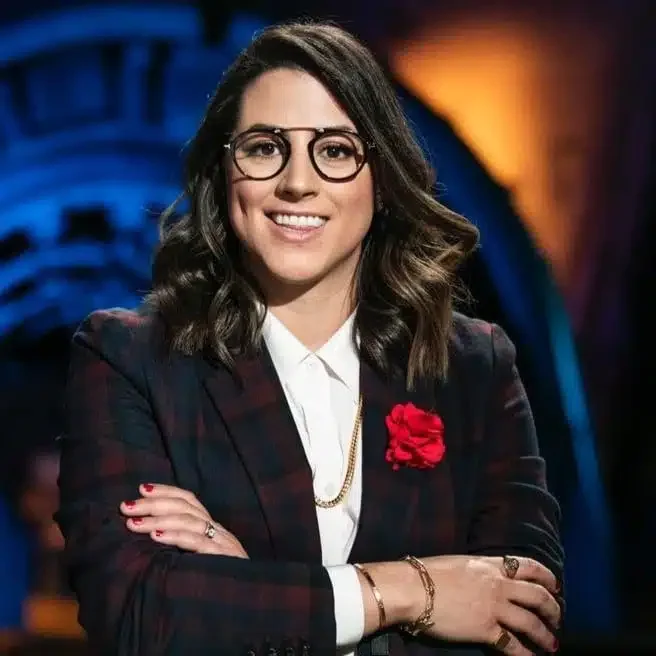 Mujer con cabello castaño, lentes redondos, vestido a cuadros y chaqueta oscura, posando con brazos cruzados y sonrisa, en un fondo oscuro con luces cálidas y frías.