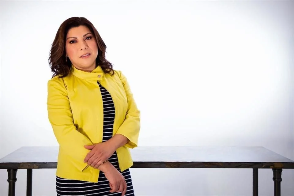 Mujer con cabello castaño oscuro, vestida con chaqueta amarilla y camiseta de rayas negras y blancas, posando con un fondo blanco y una mesa gris oscura.