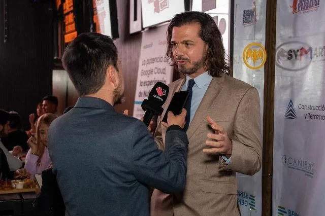 Entrevista con un hombre de cabello largo y barba a un reportero en un evento con fondos de anuncios y logotipos.