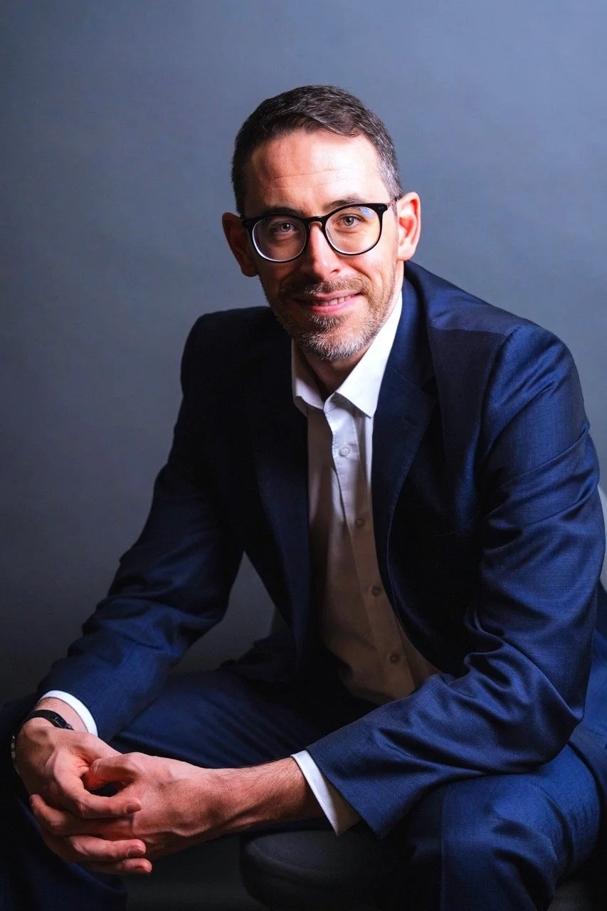 Retrato de un hombre con gafas, barba y cabello corto, vestido con un traje azul, sonriendo, frente a un fondo gris.