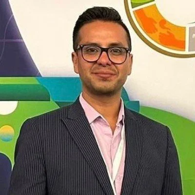 Hombre con gafas y traje en una conferencia o evento, fondo colorido con logotipos y gráficos.