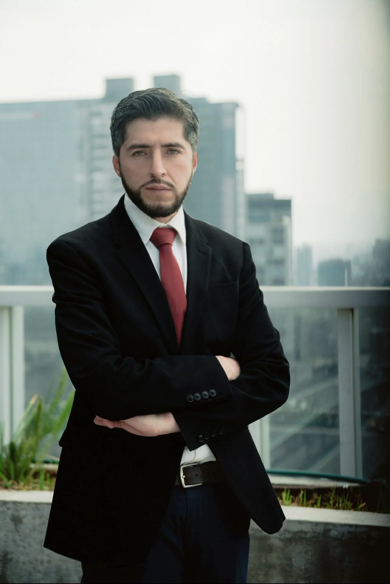 Hombre con barba y cabello oscuro, vestido con saco negro, camisa blanca y corbata roja, de pie en un balcón con fondo de edificios urbanos.