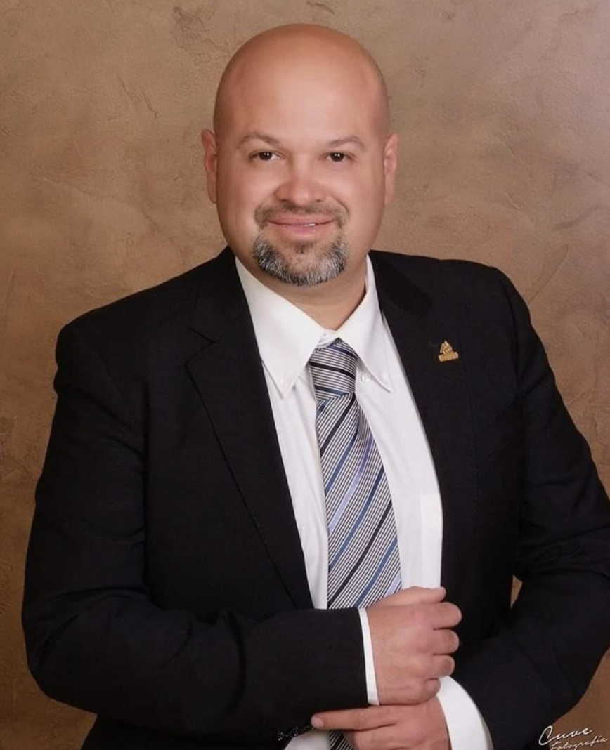 Retrato de un hombre calvo con barba, vestimenta formal con saco negro, camisa blanca y corbata a rayas, posando con fondo de pared marrón.
