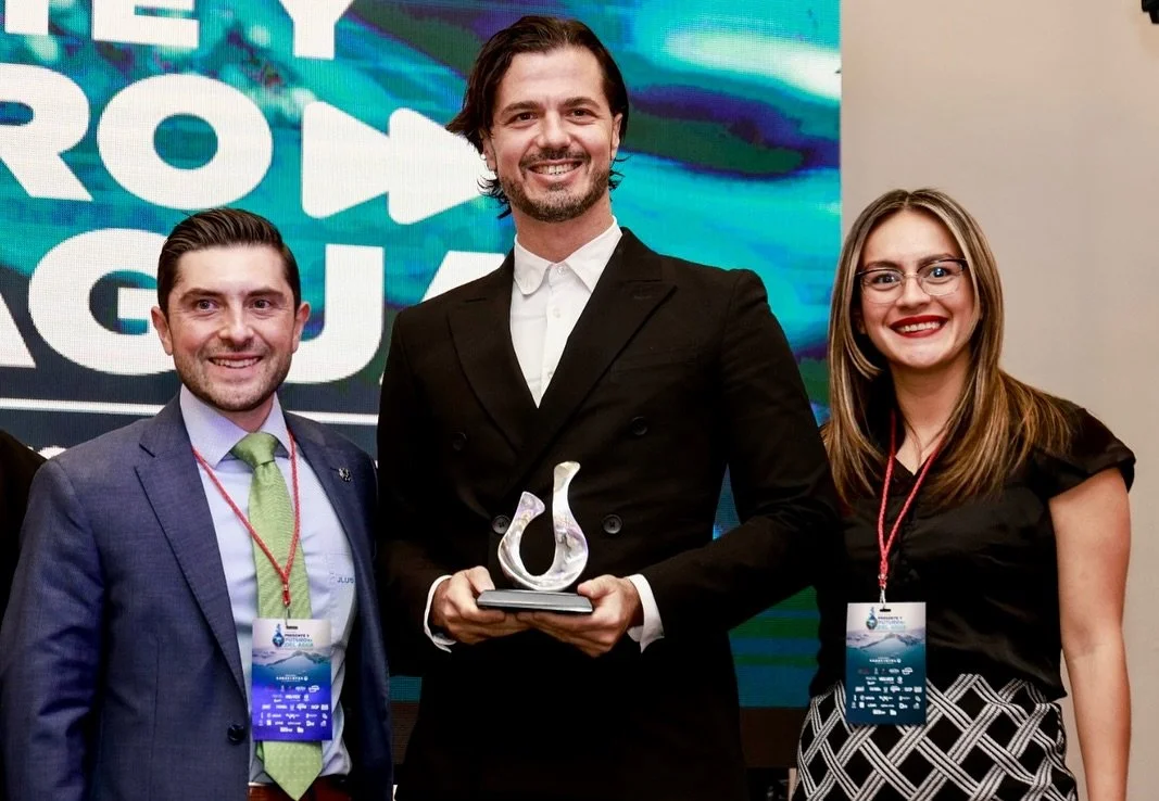 Tres personas sonriendo en un evento, una de ellas sostiene un trofeo, frente a una pantalla con palabras en inglés.