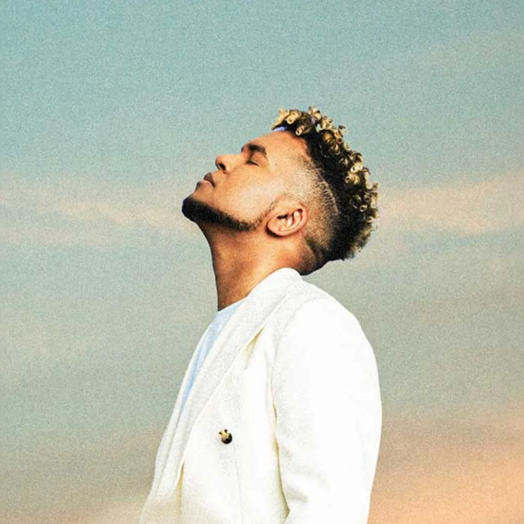 Joven con peinado de rizos cortos y rubios, usando chaqueta negra con detalles en blanco, rojo y azul, posando en un fondo rojo.