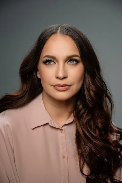 Retrato de una mujer con cabello castaño oscuro y rizado, usando blusa rosa claro, con expresión pensativa y fondo gris.