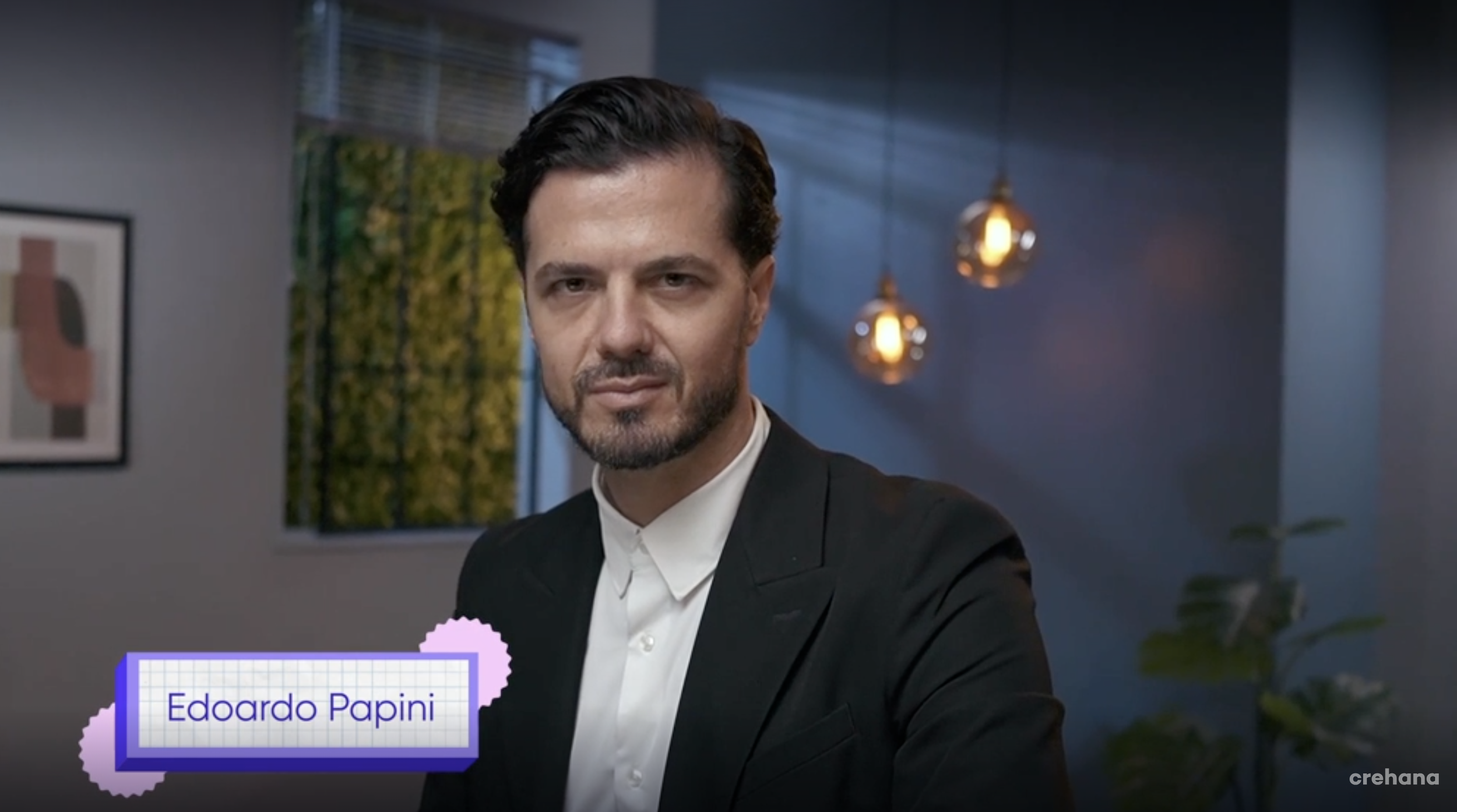 Retrato de un hombre con barba, cabello oscuro peinado hacia atrás, viste un saco negro y camisa blanca, en un ambiente moderno con iluminación cálida y plantas decorativas.