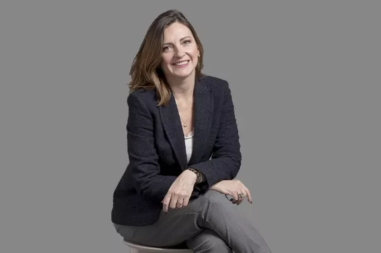 Mujer sonriente con cabello castaño, vestida con saco oscuro y pantalones grises, sentada sobre una silla gris, fondo gris.