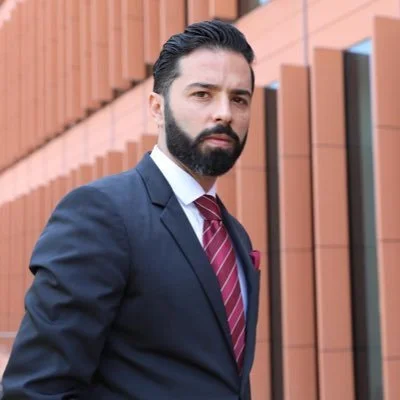 Hombre con barba y barba, vestido con traje azul marino y corbata a rayas, de fondo con paredes de madera.
