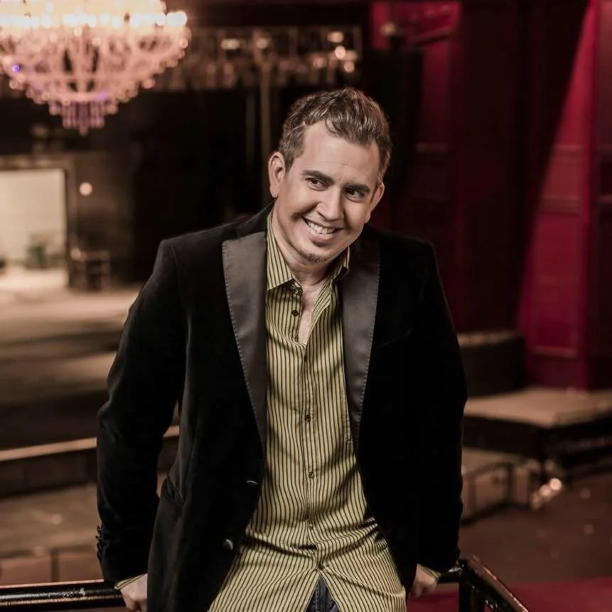 Hombre sonriendo, sentado en una barandilla en un teatro con candelabro y cortinas rojas en el fondo.