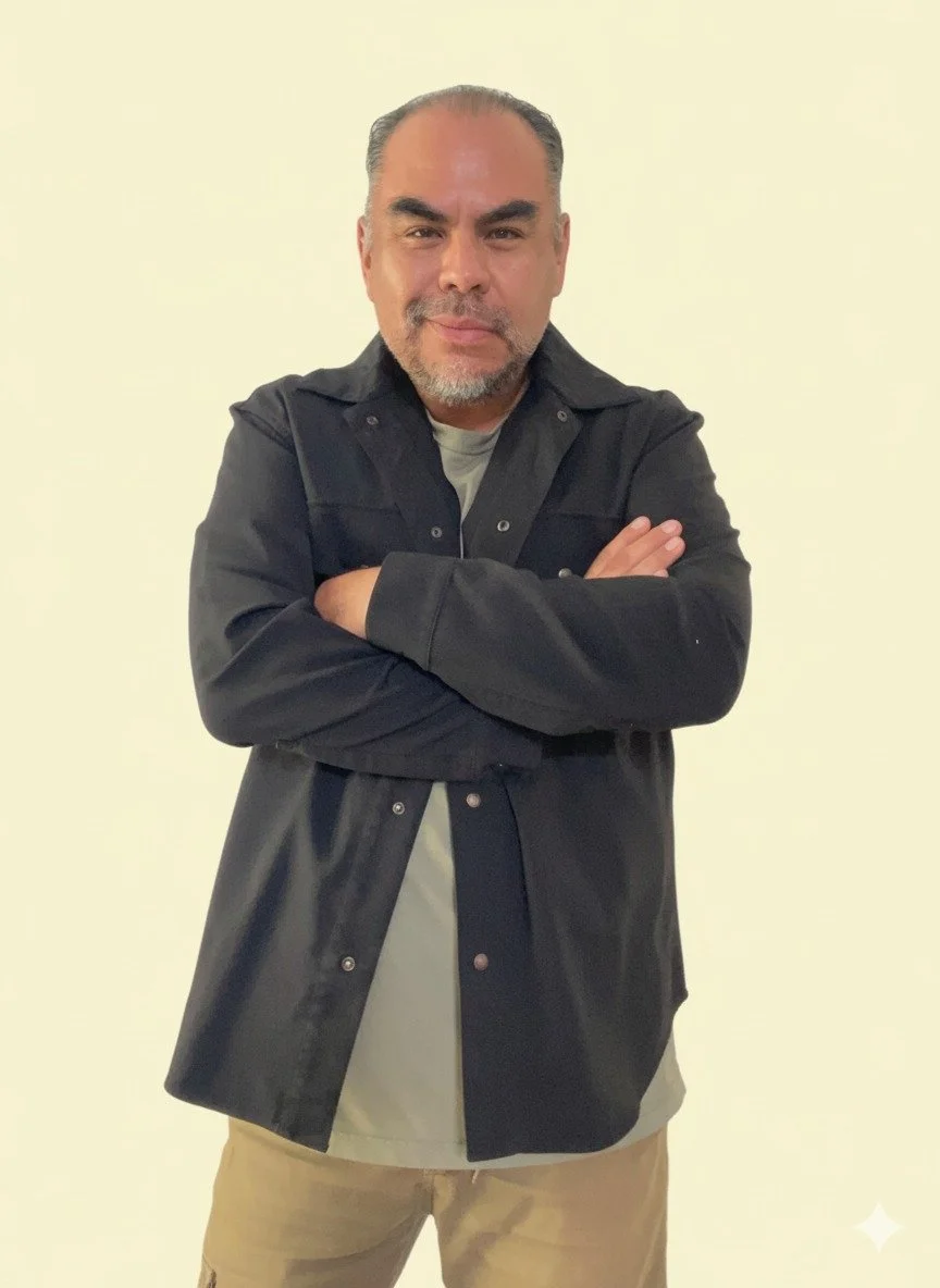 Hombre con barba y cabello corto, vestimenta casual oscura, cruzado de brazos, sonriendo ligeramente, fondo blanco.