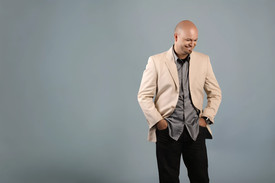 Hombre con barba, sonriente, vestido con saco oscuro y camisa blanca, en un fondo negro.