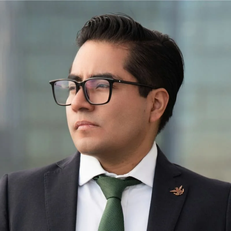 Hombre vestido con traje y corbata, usando gafas, mirando pensativamente hacia un lado.