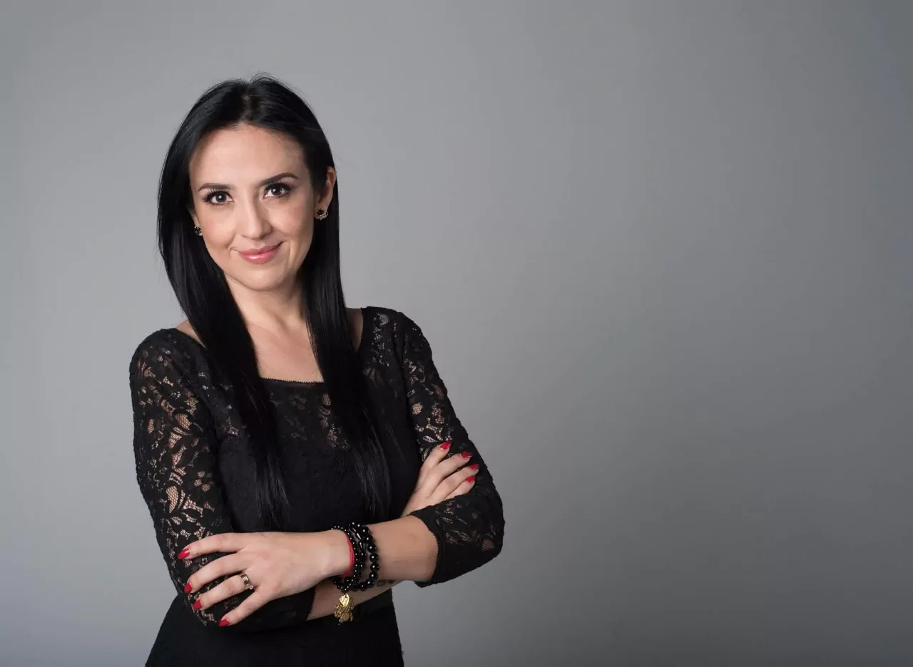 Mujer con cabello negro, vestido negro de encaje, con pulseras y aretes, sonrisa y brazos cruzados frente a fondo gris.