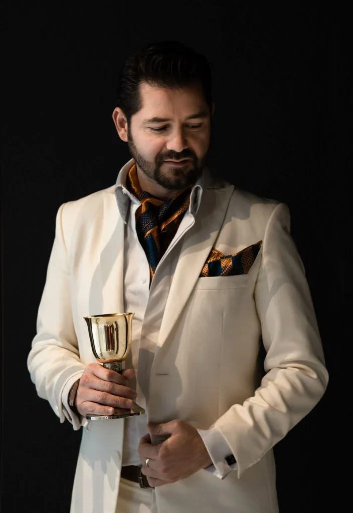 Hombre con traje blanco y pañuelo de bolsillo, sosteniendo una copa dorada, con fondo negro.