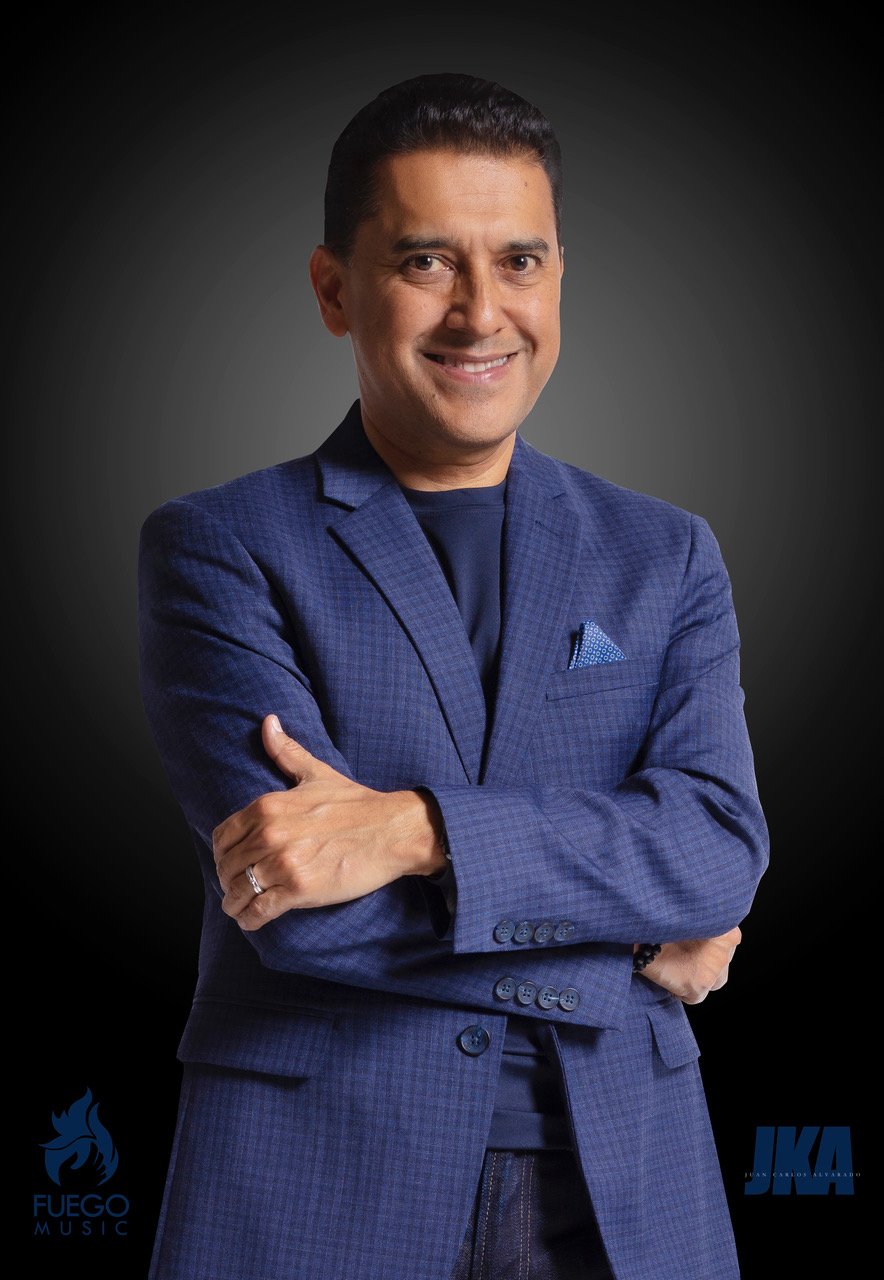 Hombre con traje azul, sonrisa, brazos cruzados, fondo oscuro, logotipos en las esquinas inferiores izquierda y derecha.