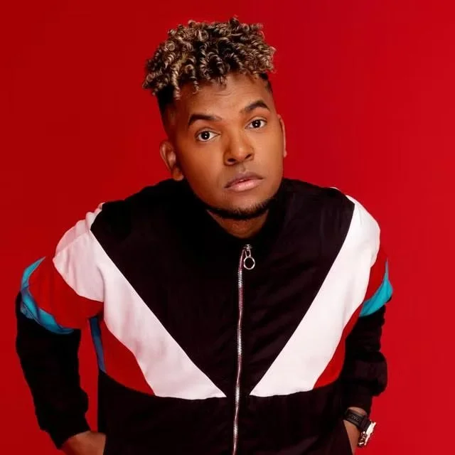 Joven con cabello rizado y expresión neutral, viste una chaqueta de colores rojo, blanco, negro y azul, y está de pie frente a un fondo rojo.