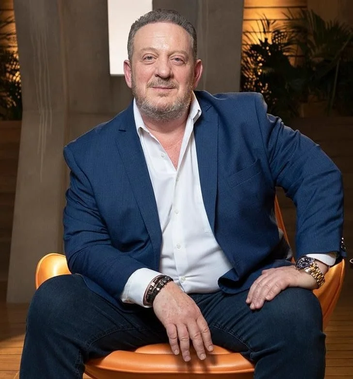 Retrato de un hombre de cabello castaño claro, con barba y expresión seria, usando una chaqueta negra y una camisa blanca, con un reloj dorado en la muñeca izquierda, en un fondo oscuro.