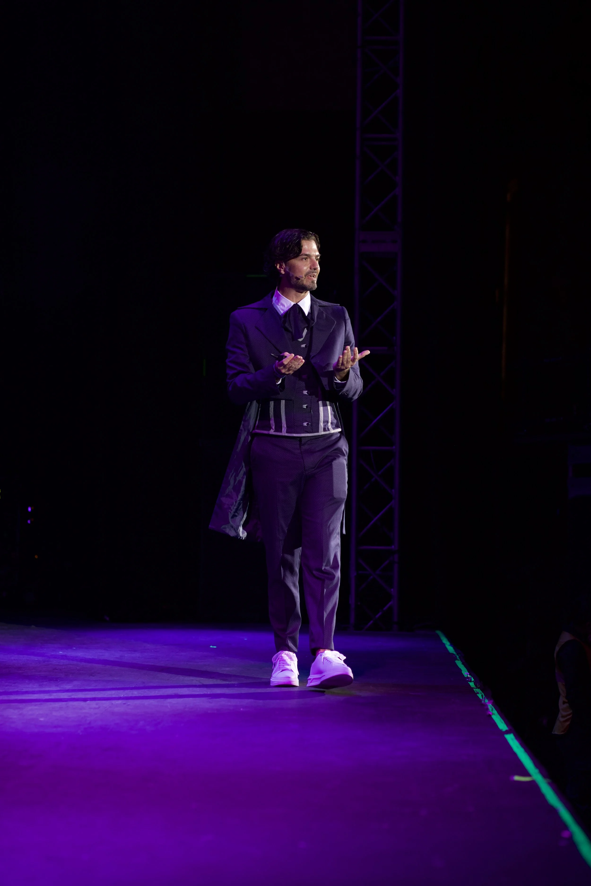 Hombre en un escenario con iluminación morada, vestido con ropa formal y haciendo una presentación o discurso.