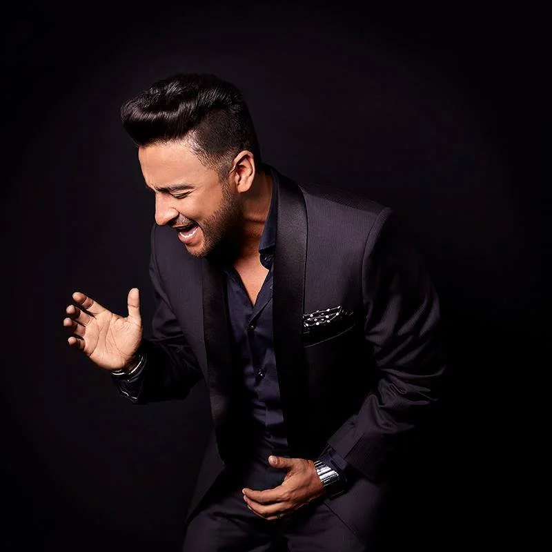 Hombre con barba, gafas de sol grandes, chaqueta de cuero negra y cabello peinado hacia atrás, posando con las manos cruzadas en un fondo de pared de ladrillo y cerca de una cerca de alambre.