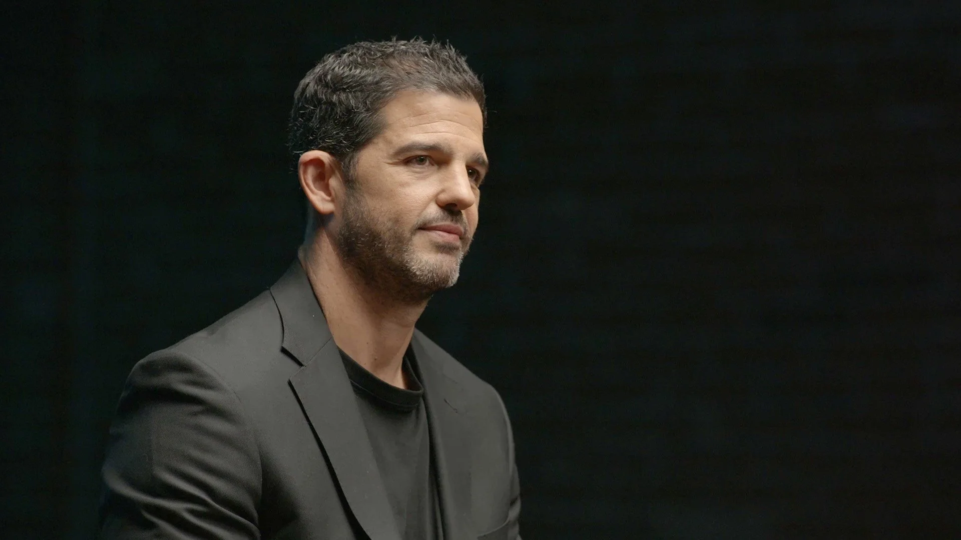 Hombre con barba y cabello corto, viste un saco negro y camiseta negra, mira hacia la izquierda, fondo negro.