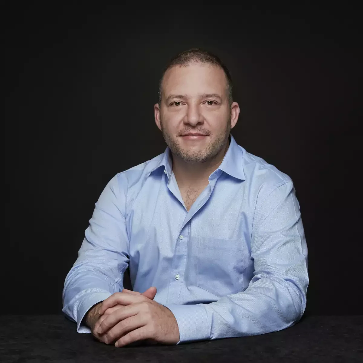 Retrato de un hombre con camisa azul claro, sonriendo, con fondo negro.