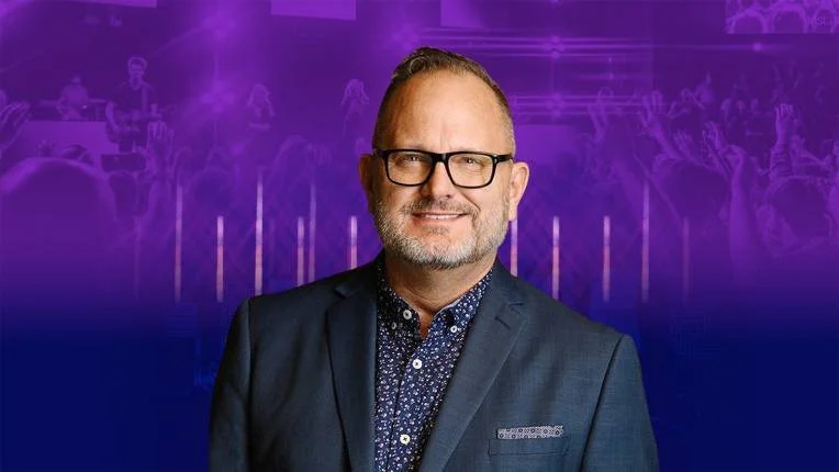 Hombre con gafas y barba, usando un traje oscuro, en un escenario con fondo morado y azul con siluetas de personas y luces.