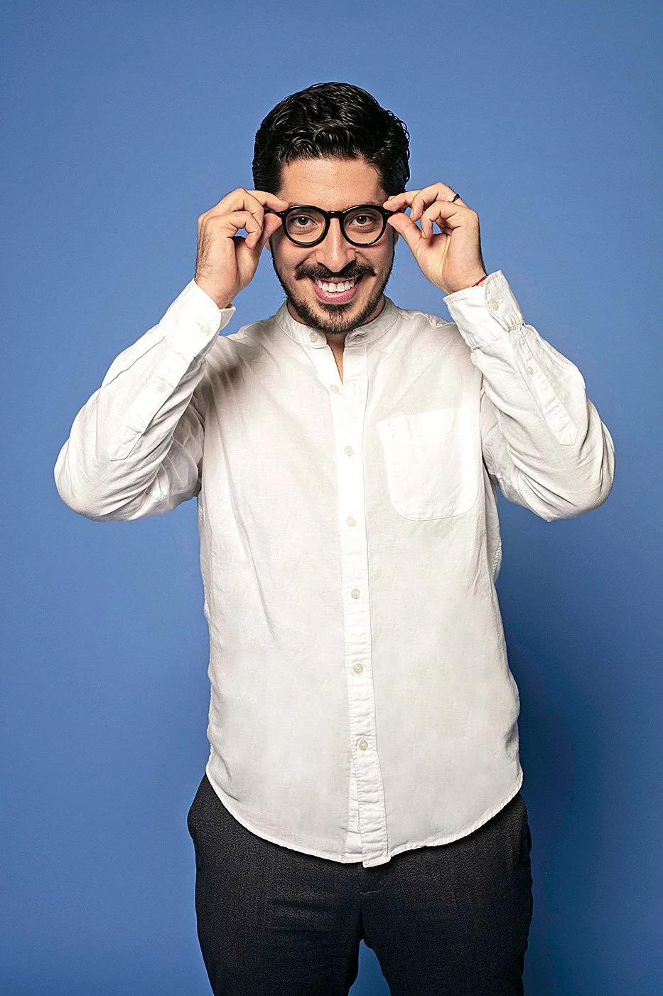 Hombre sonriendo, usando gafas y camisa blanca, ajustando sus gafas, frente a un fondo azul.