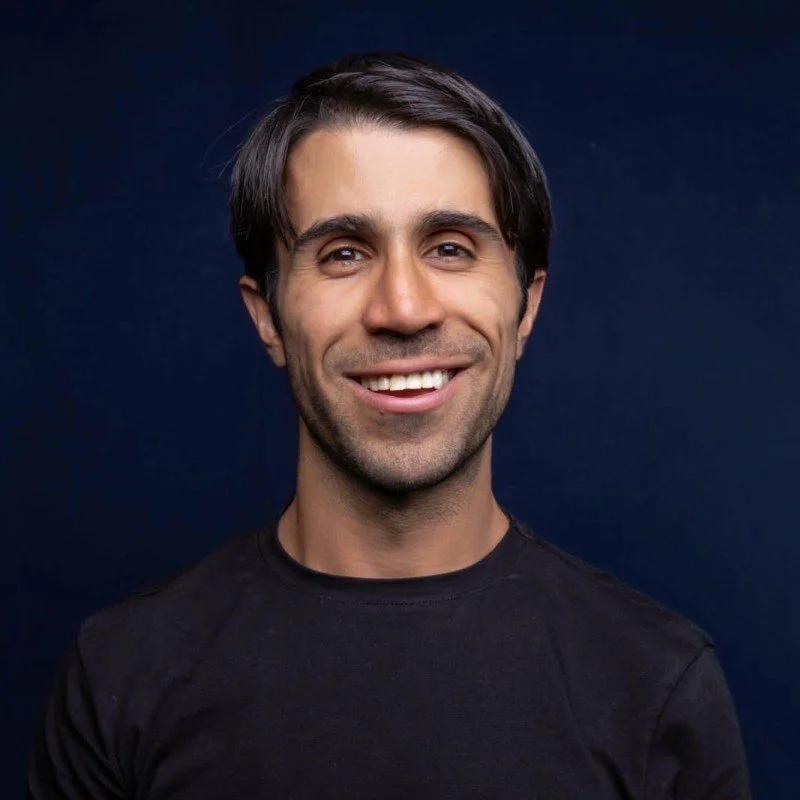 Retrato de un hombre sonriendo, con cabello oscuro y camiseta negra, fondo azul oscuro.