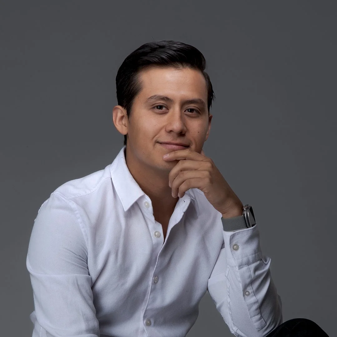 Joven hombre con cabello oscuro peinado hacia atrás, sonriendo ligeramente, usando camisa blanca y reloj, con fondo gris