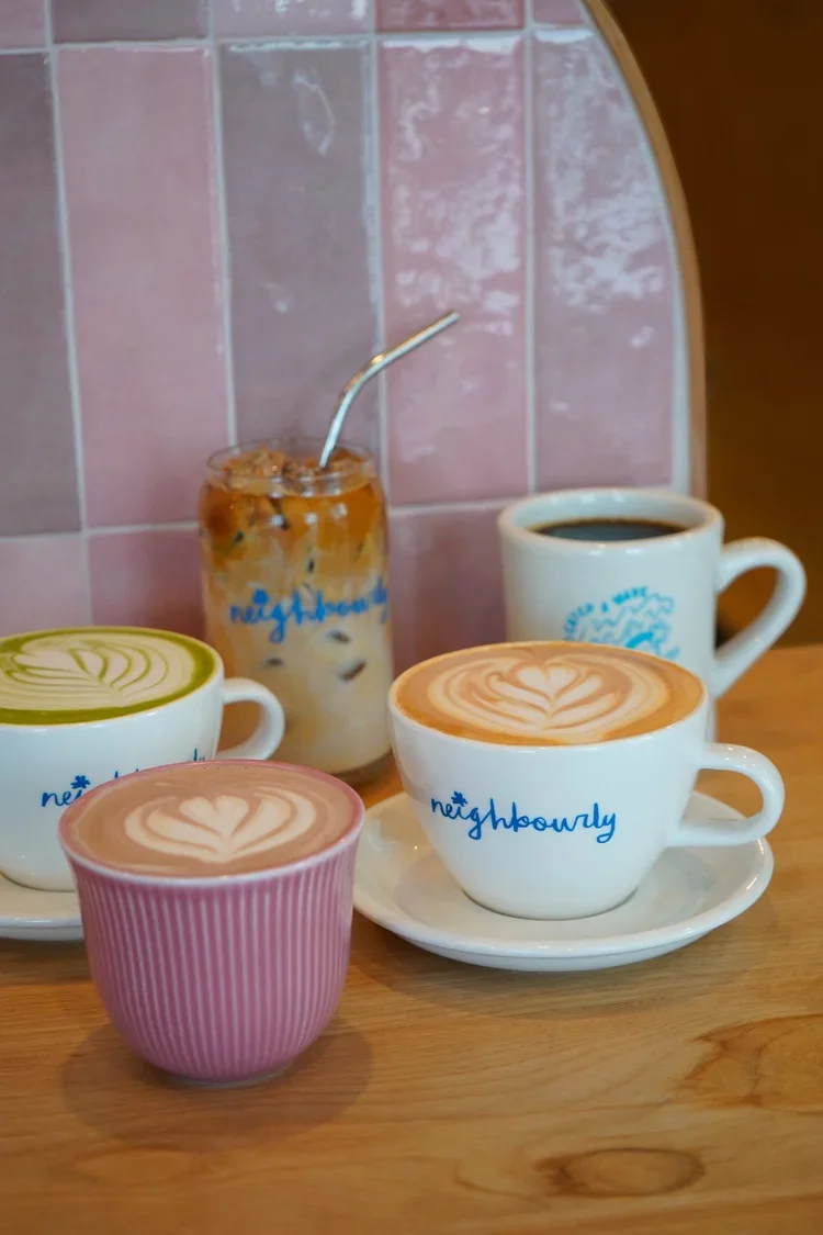 Four coffee drinks with latte art sit on a wooden table in front of a pink tiled wall. Two are in branded white cups with saucers, one in a pink striped cup, and one in a plain white mug. A tall glass of iced coffee with a straw is in the background.