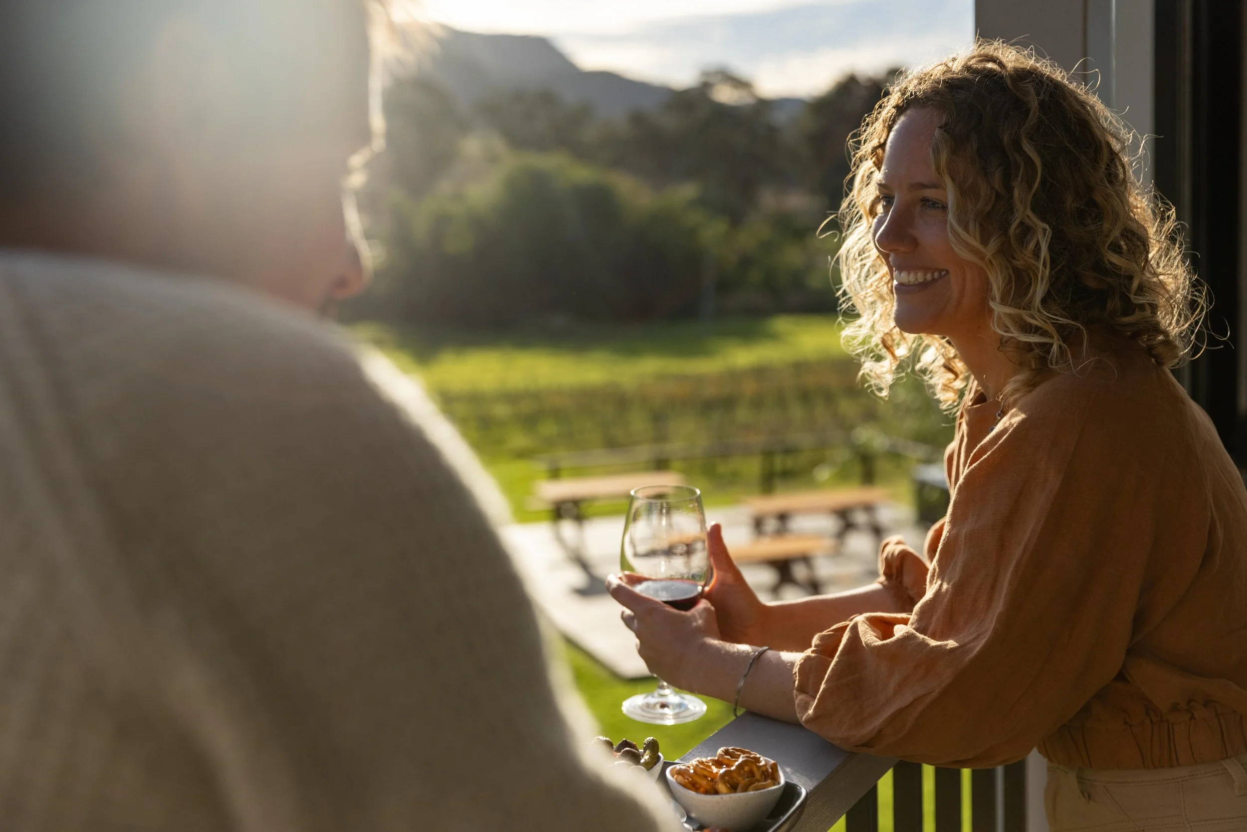 A woman with curly blonde hair smiling and holding a glass of red wine, talking to a person whose back is visible, on a balcony with a green outdoor landscape in the background.