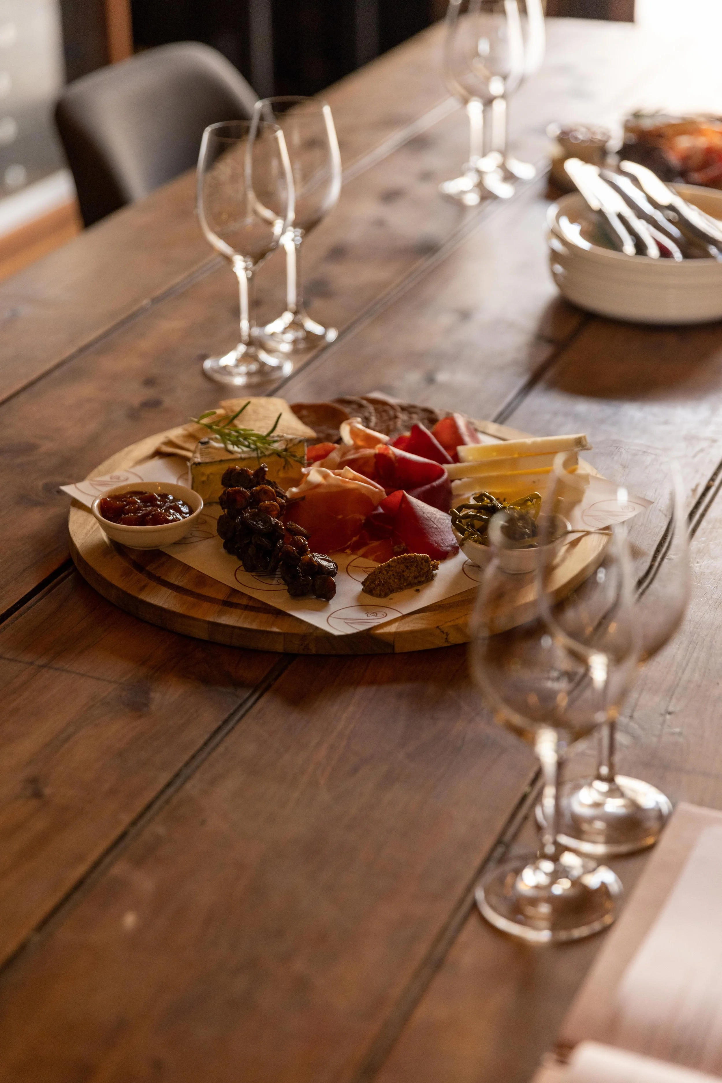 Charcuterie board with assorted cheeses, meats, grapes, and a small bowl of jam on a wooden table near empty wine glasses.