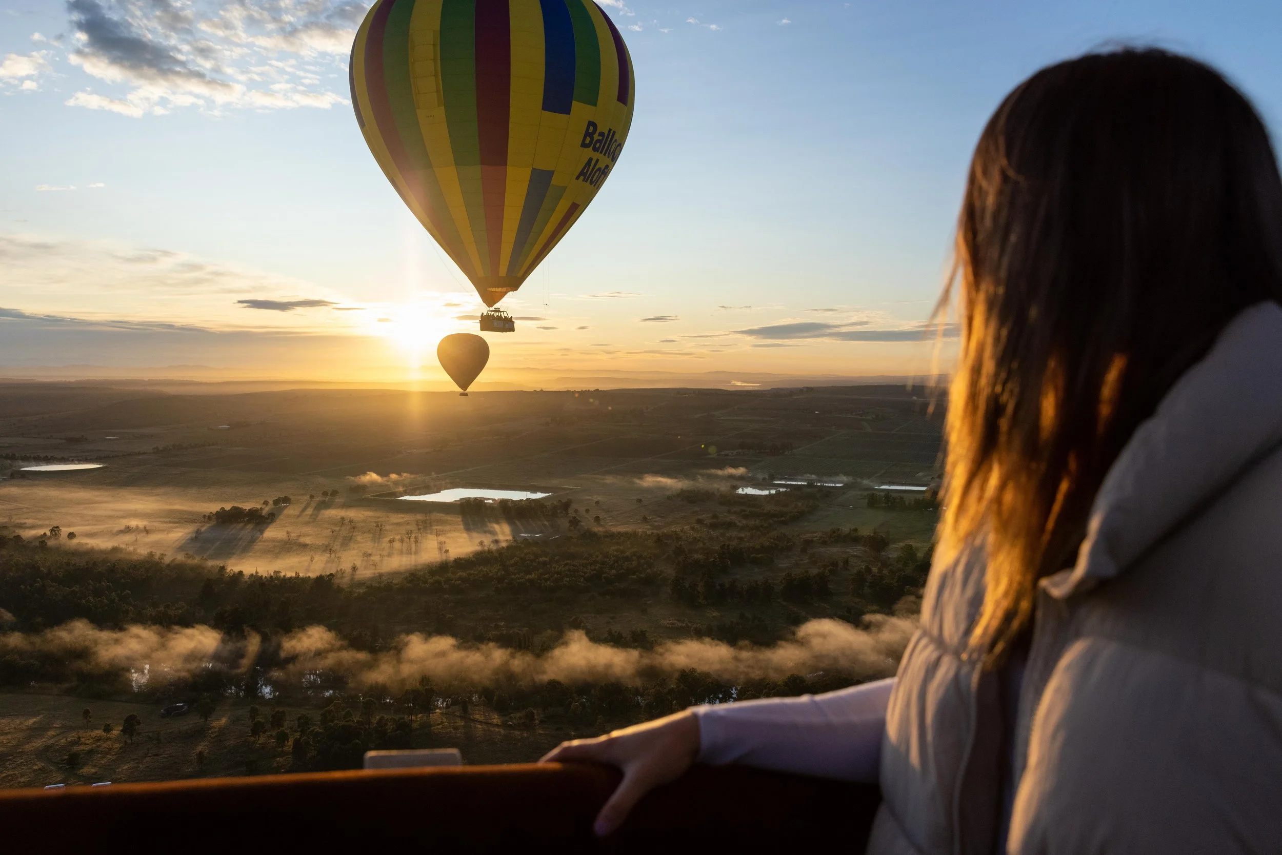 A woman gazing at hot air balloons flying during sunrise over a landscape of fields and lakes.