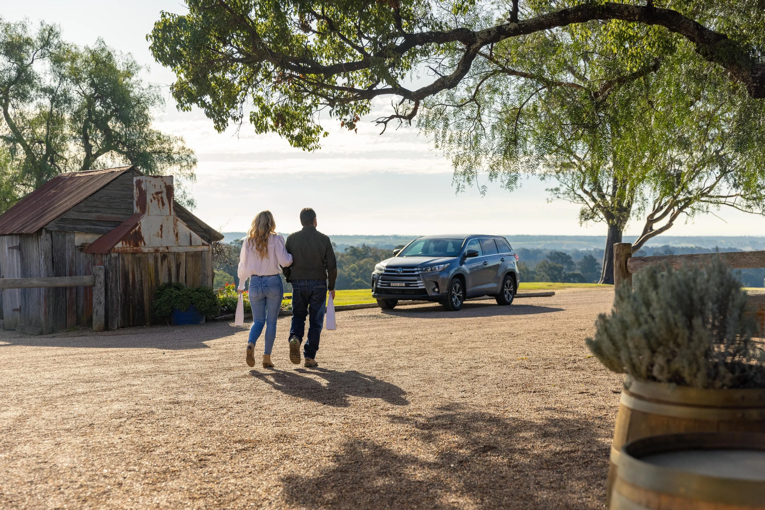 Couple walking away from a parked gray SUV in a rural landscape with trees, a rustic shed, and rolling hills in the background during daylight.