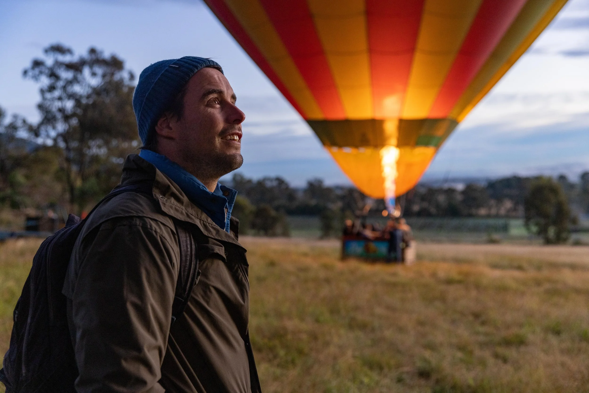 Man in jacket and beanie watching a hot air balloon being inflated at sunrise or sunset in a rural field.