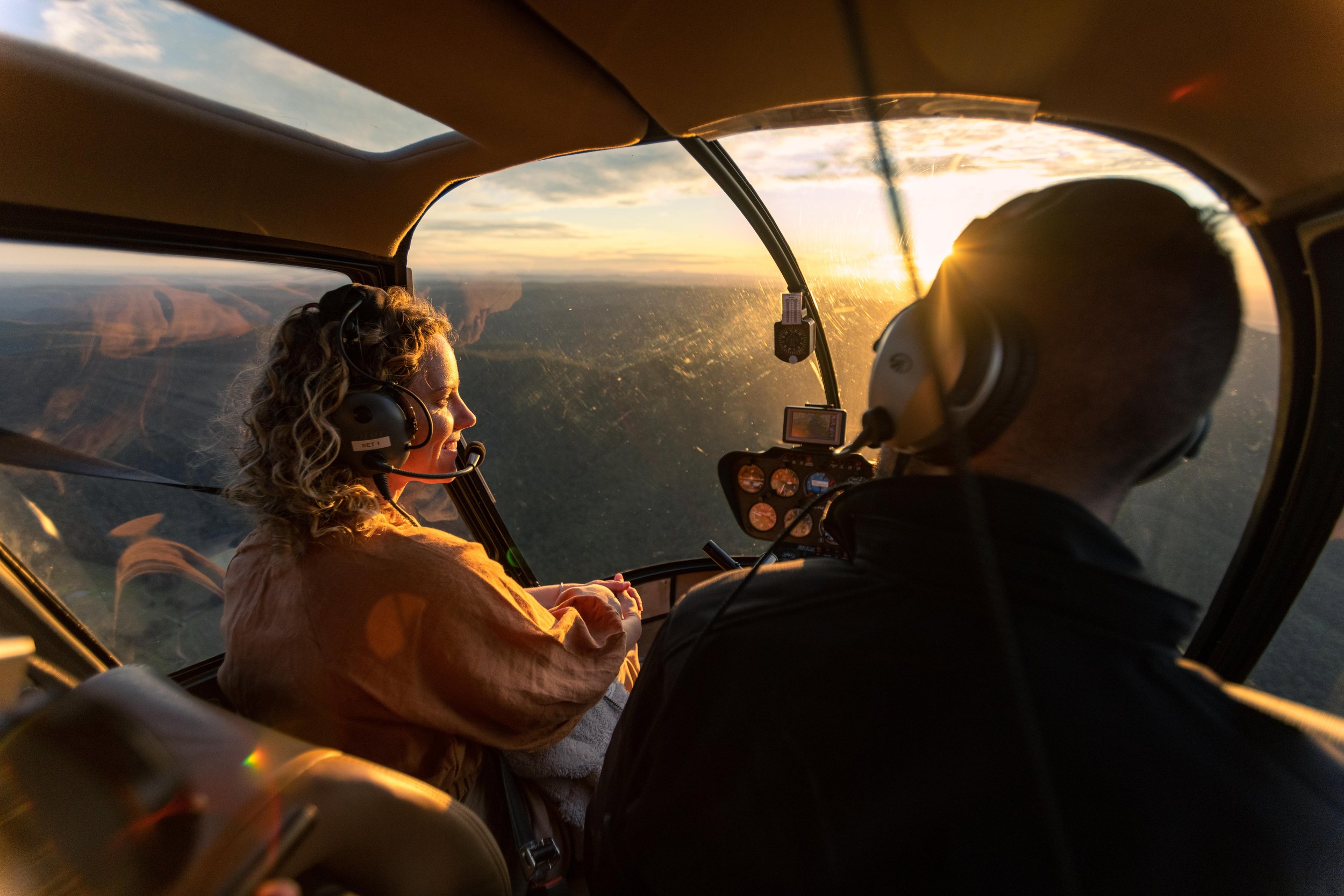 Pilot and passenger in a helicopter flying at sunset over a landscape