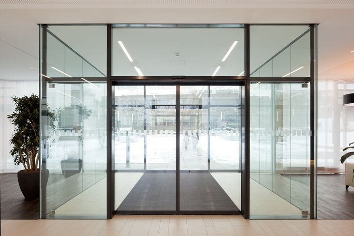 Glass entrance door with sliding panels, flanked by glass walls, in a modern building lobby with potted plant on the left and seating area on the right.
