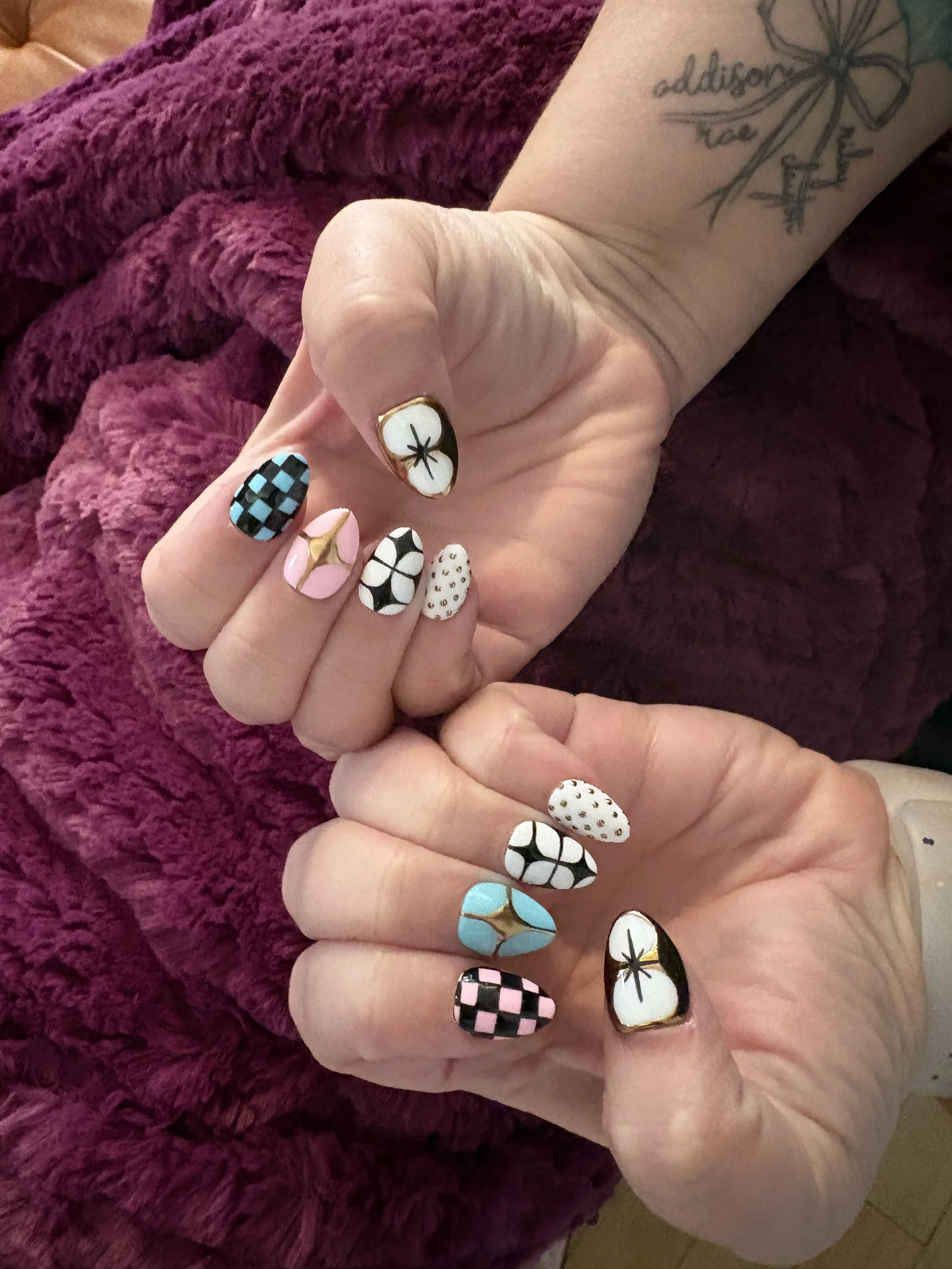 Close-up of hands with colorful, patterned fingernails, resting on a purple fuzzy blanket. Some nails feature checkered, floral, polka dot, and geometric designs.