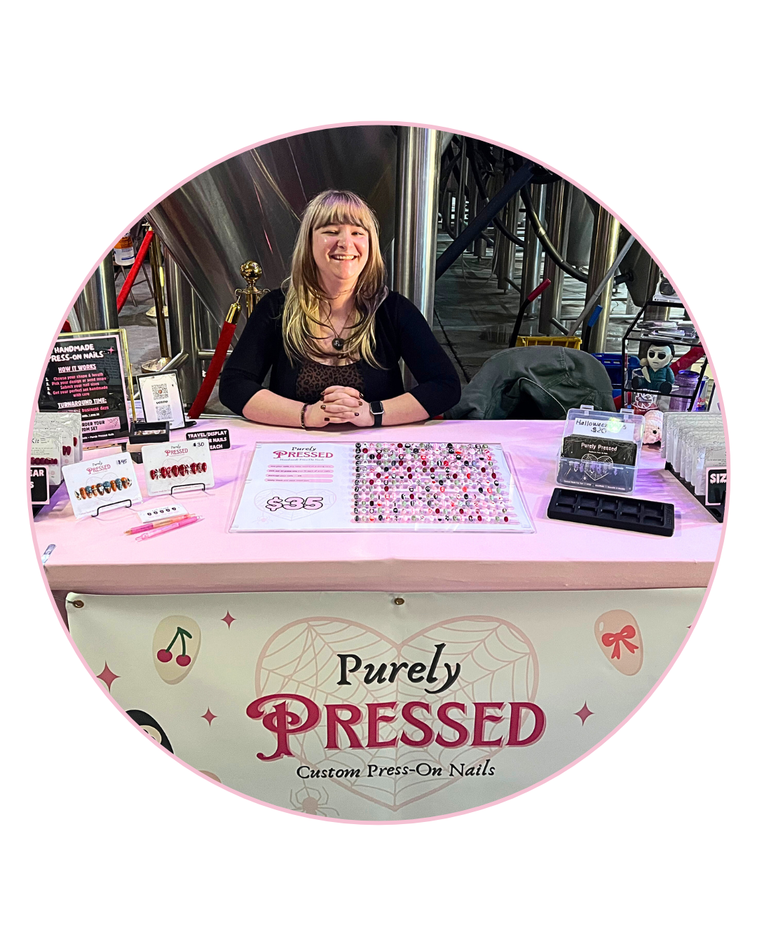 A woman sitting at a booth selling custom press-on nails with various nail designs, decorated with pink and Halloween-themed items at an event.