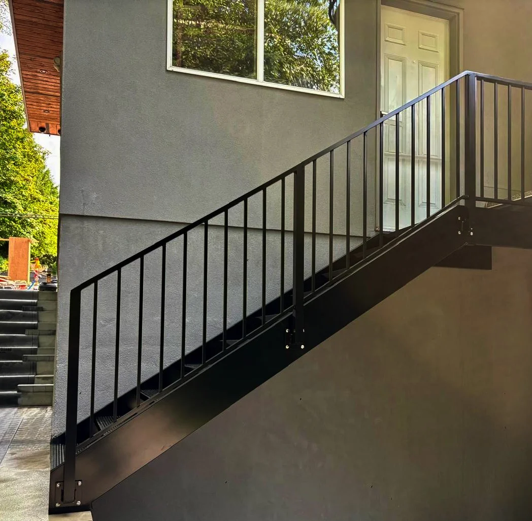 Modern outdoor steel stair railing featuring clean vertical balusters and a black metal handrail leading to a residential entrance.