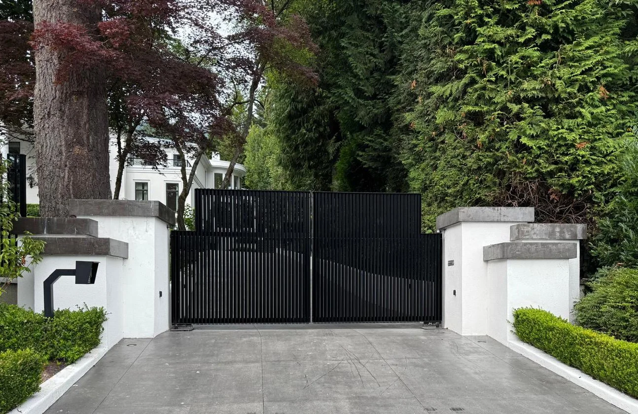 Black sliding gate with white pillars and lush green trees and shrubbery in the background.