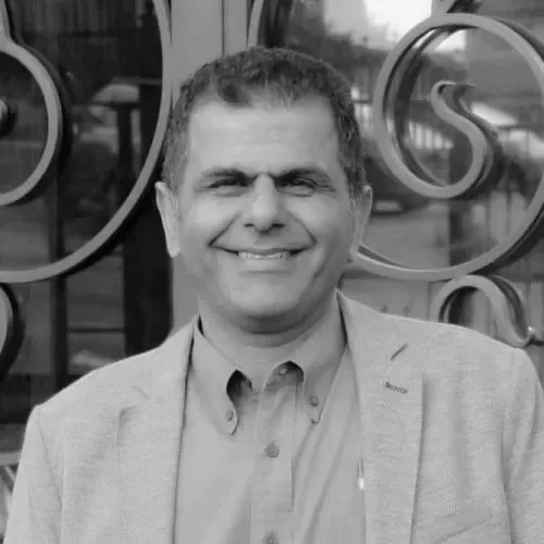 Smiling man in light gray blazer and shirt standing outdoors in front of a glass window with decorative metal design.