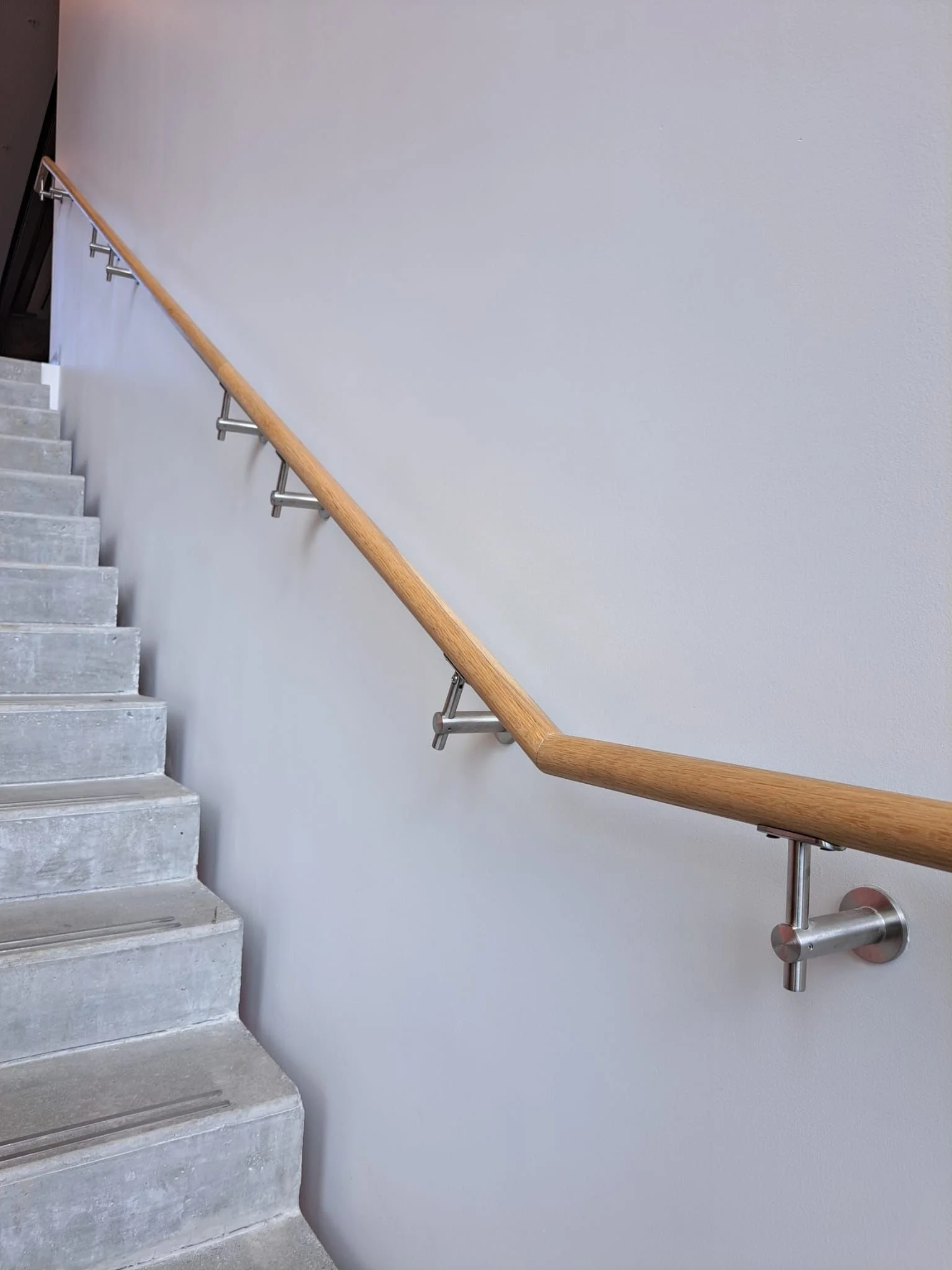 Concrete stairs with a wooden handrail and metal supports, next to a plain light-colored wall.