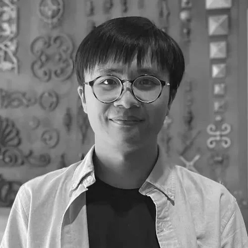 Young person with short black hair and glasses, wearing a light blue shirt and black t-shirt, smiling in front of a decorative gray wall with various patterns.