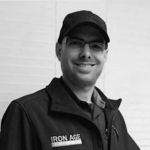 A smiling man wearing glasses, a black baseball cap, and a black jacket with 'Iron Age' embroidered on it, standing against a white tiled wall.