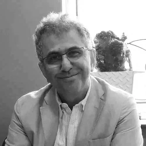 A man with gray curly hair and glasses smiling, wearing a beige blazer and a white shirt, sitting indoors near a window with plants in the background.