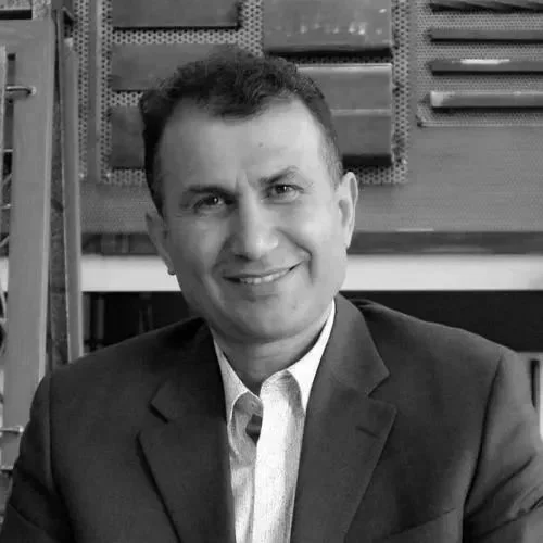 A man wearing a dark suit and white shirt, smiling, with a industrial or workshop background.